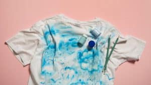Camiseta branca com manchas azuis em fundo rosa, representando roupas sujas de crianças.