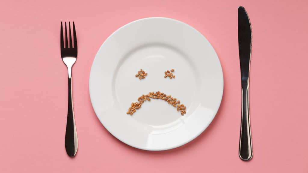 Prato com grãos formando uma carinha triste, acompanhado por garfo e faca em um fundo rosa, simbolizando a alergia alimentar.