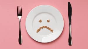 Prato com grãos formando uma carinha triste, acompanhado por garfo e faca em um fundo rosa, simbolizando a alergia alimentar.