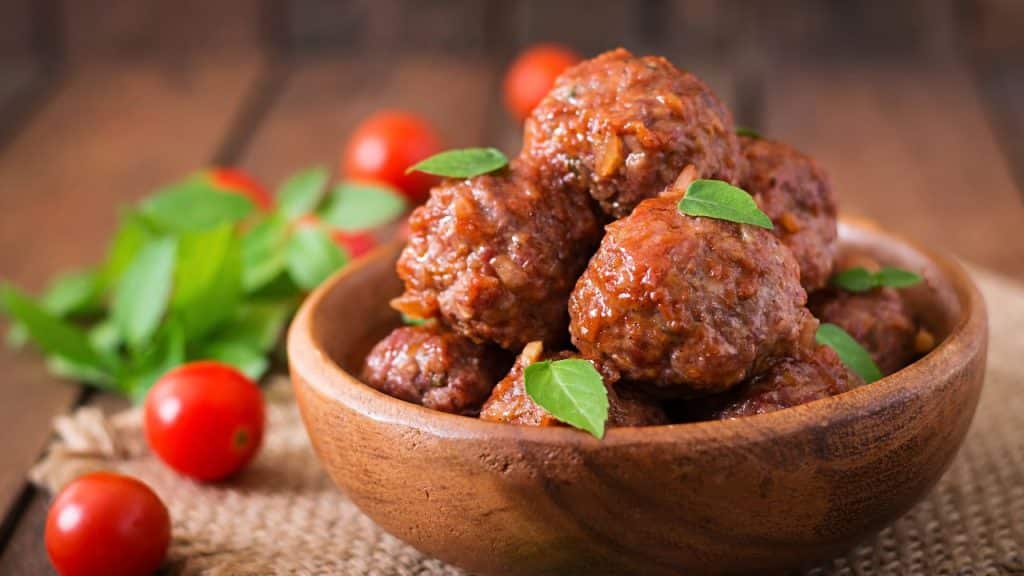 Almôndega ao molho de tomate em uma tigela de madeira com tomate cereja atrás.