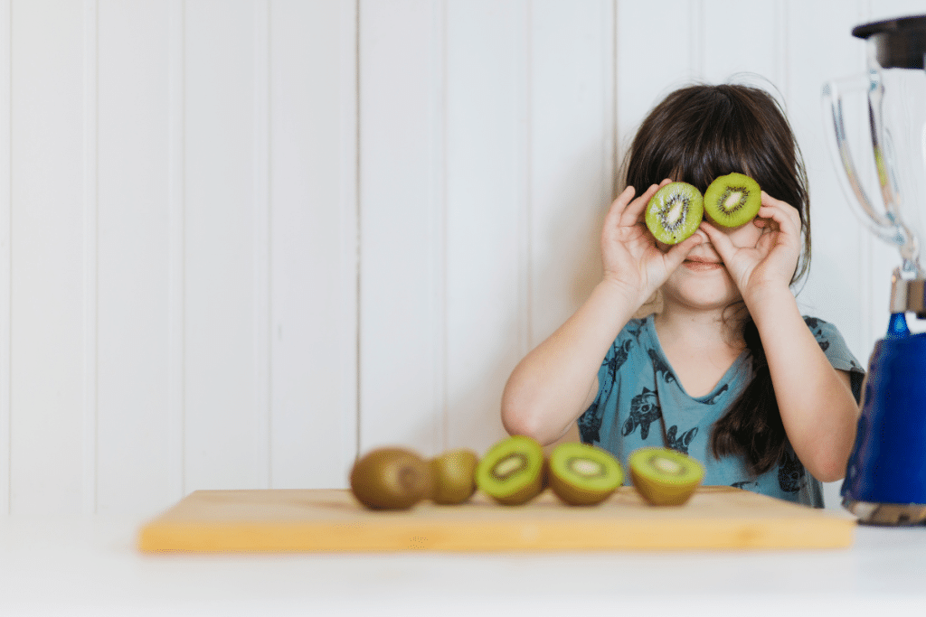 Menina segurando dois kiwis na frente dos olhos e na sua frente também tem alguns cortados.