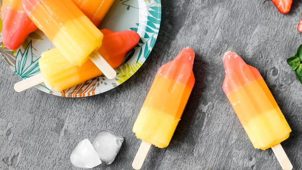 Dois picolés de salada de fruta em cima de um fundo de madeira cinza com um prato de picolés ao lado.