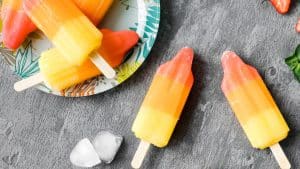 Dois picolés de salada de fruta em cima de um fundo de madeira cinza com um prato de picolés ao lado.