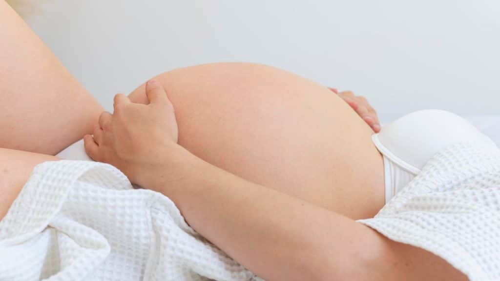 Close na barriga de uma mulher grávida vestindo branco, simbolizando o parto.