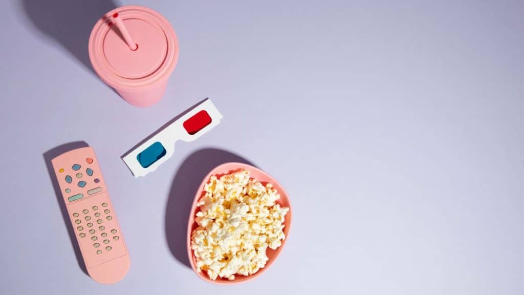 Balde de pipoca, controle remoto, copo e óculos 3D em tons rosados representando filmes da Pixar.