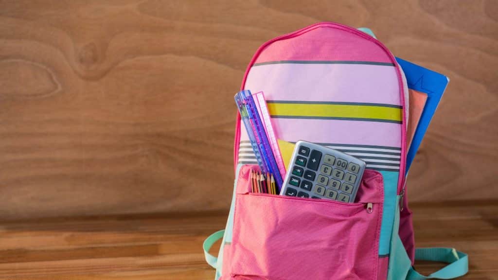 Mochila rosa cheia de materiais de escola em um fundo de madeira.