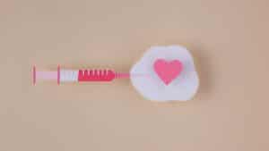Seringa de vacinação rosa junto a um algodão com coração rosa sobre um fundo da mesma cor.