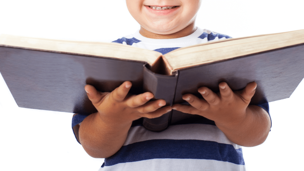 Criança com um livro grande na mãe e sorrindo. Na imagem, ela está em um fundo branco