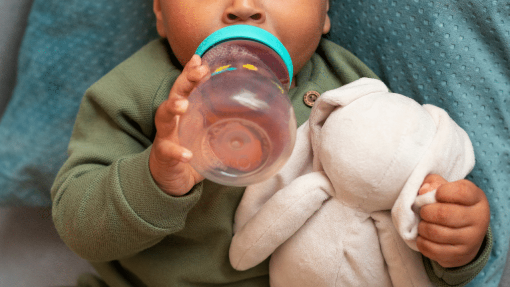 Um bebê segurando uma mamadeira de água e agarrrado em um ursinho