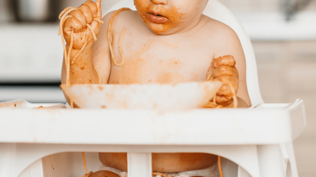 Criança comendo macarrão, enquanto está com as mãos sujas e o babador também