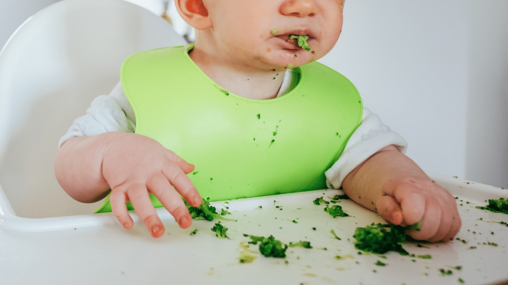 Criança comendo, provavelmente um=algum alimento que seja folha. Ela está com um babador verde e com a boca suja