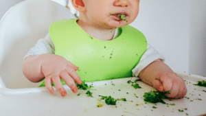 Criança comendo, provavelmente um=algum alimento que seja folha. Ela está com um babador verde e com a boca suja