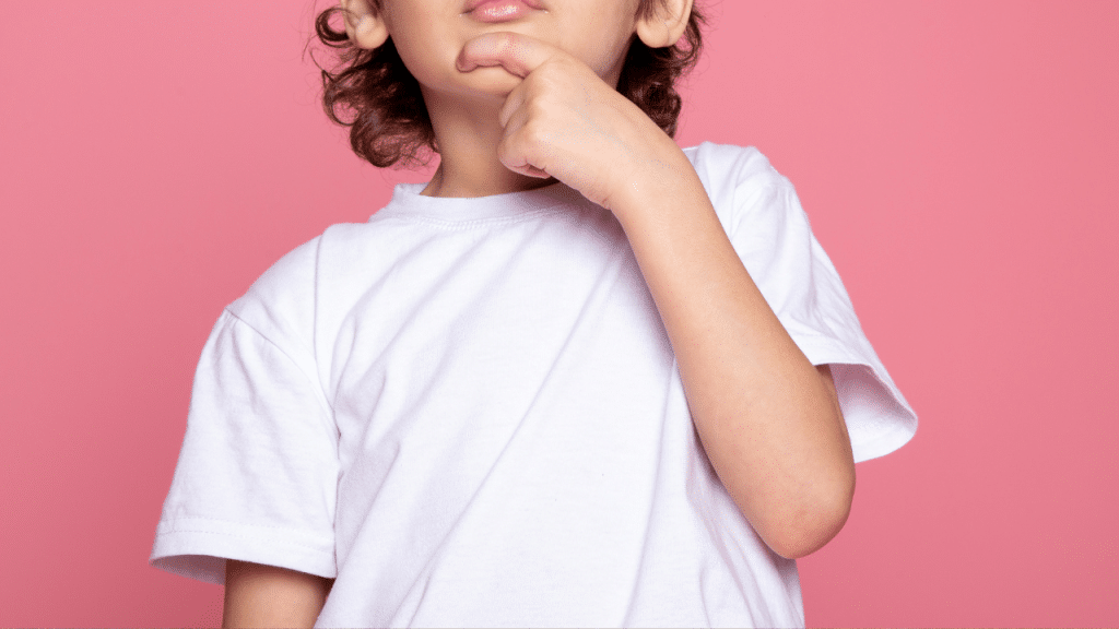 Criança com a mão ma boca, uma camisa branca e um fundo rosa.