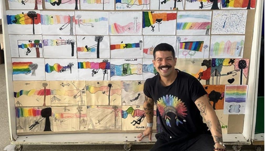 MENA agachado em frente a um mural com desenhos de crianças.