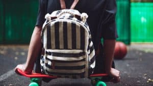 Jovem sentado num skate com uma mochila maluca nas costas.