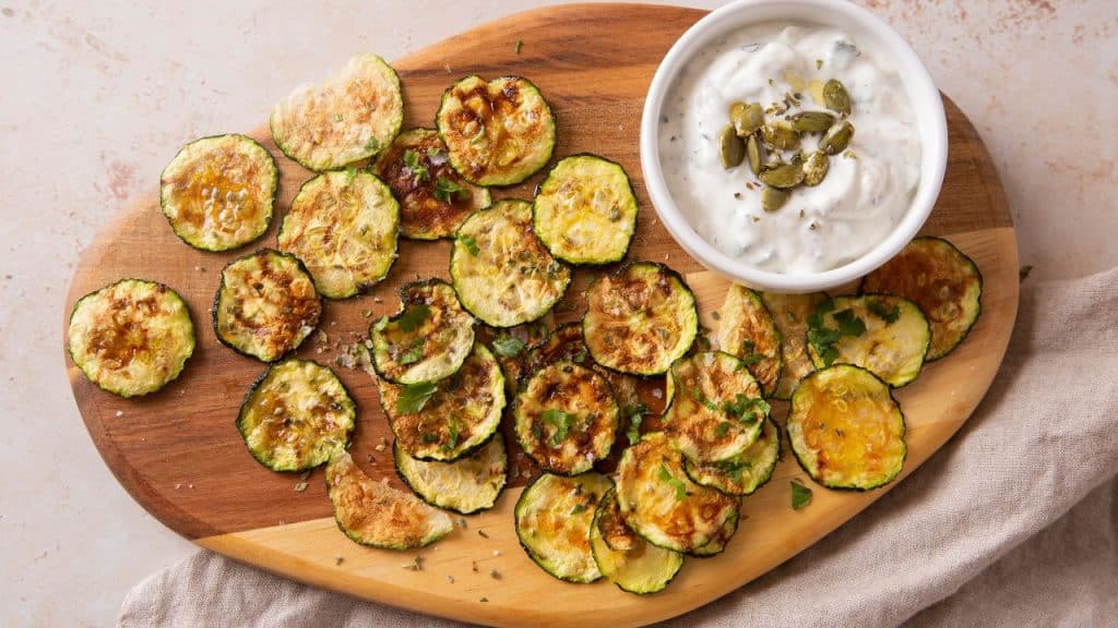 Chips de abobrinha em tábua de madeira com molho branco ao lado.