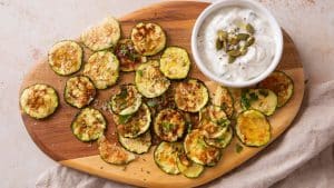 Chips de abobrinha em tábua de madeira com molho branco ao lado.