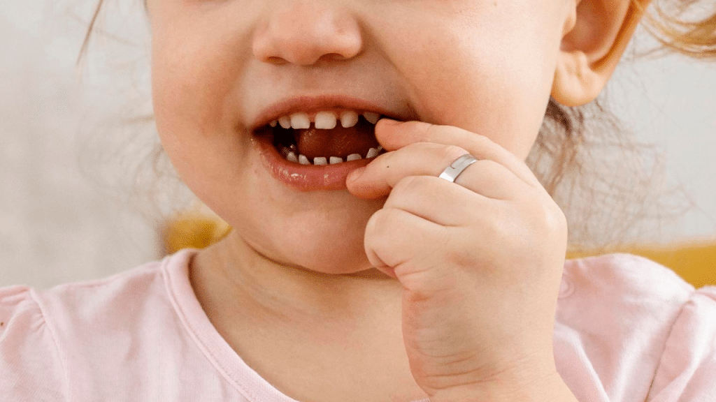 Criança com dedo na boca, dentes aparentes e camiseta rosa