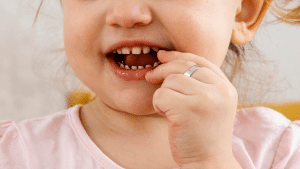 Criança com dedo na boca, dentes aparentes e camiseta rosa